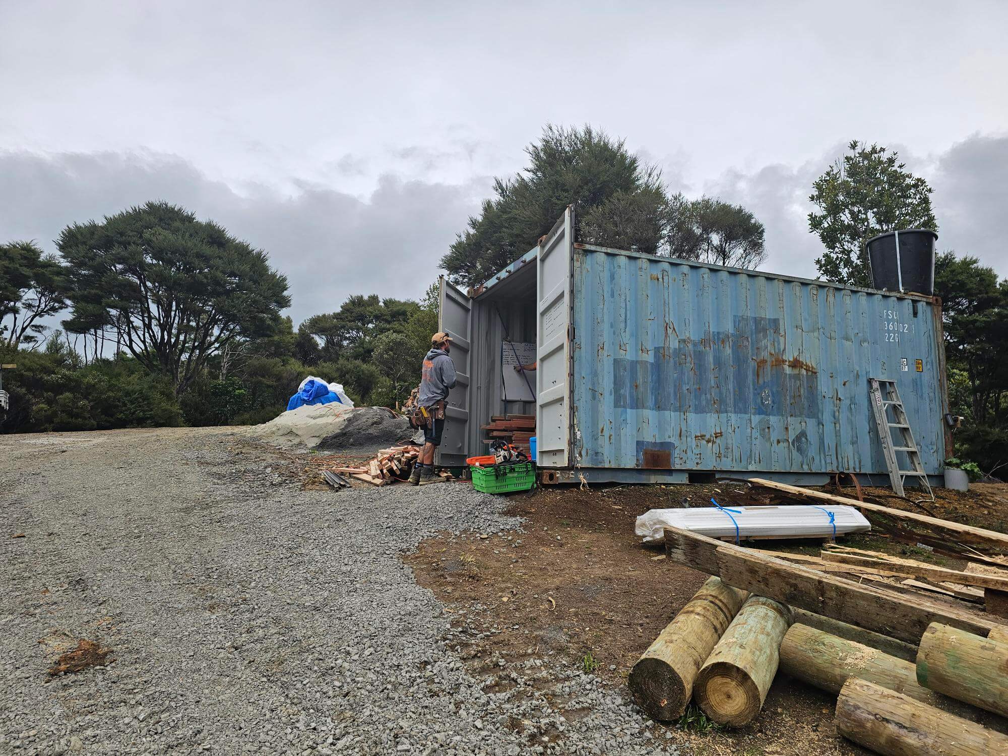 A person stands near an open shipping container on a gravel path, surrounded by logs and construction materials. The scene is set outdoors with trees and overcast skies in the background.
