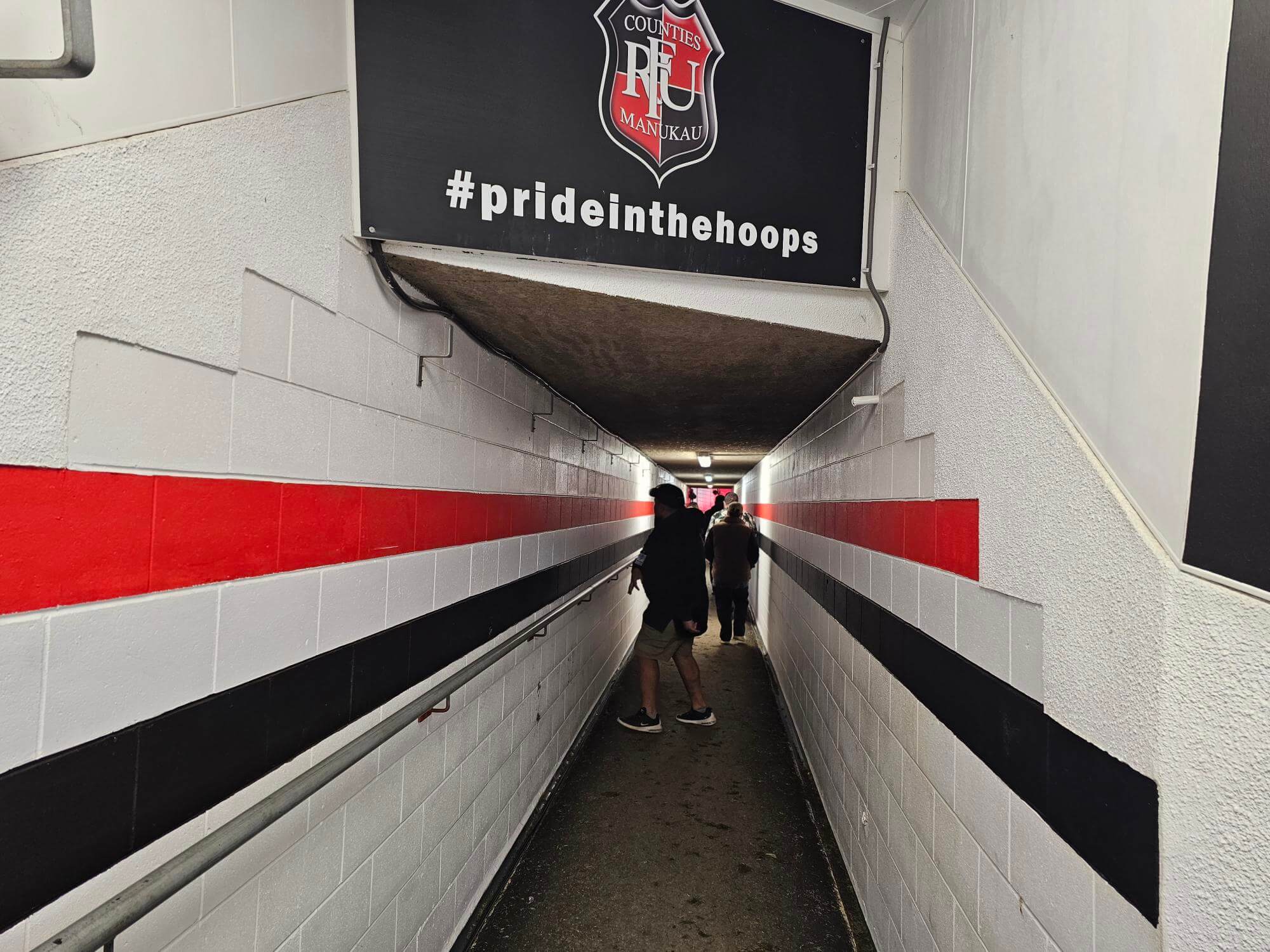 A narrow sports tunnel with white walls, featuring red and black horizontal stripes. A few people walk towards the exit. The overhead sign reads pride in the hoops, along with a sports team emblem.