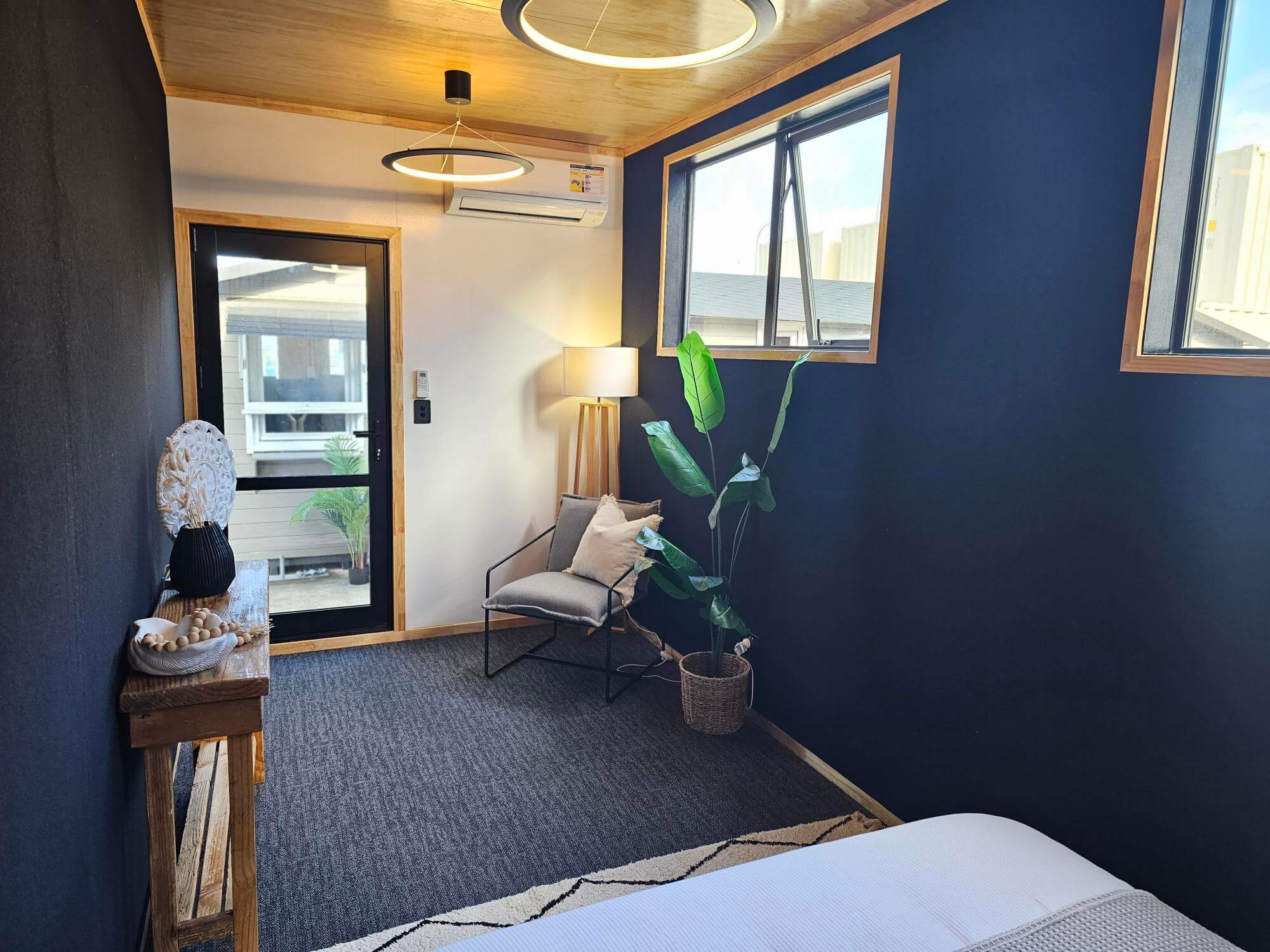 Modern bedroom with dark accent wall and wooden ceiling. Features a bed partially in view, armchair, potted plant, and a floor lamp beside a window. A console table with decorative items and a glass door leading outside completes the cozy setup.