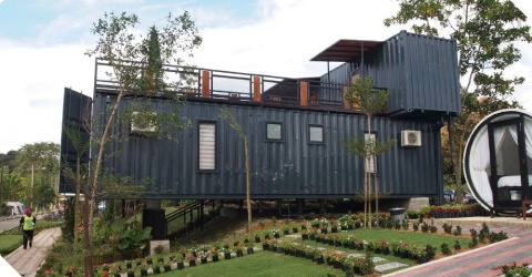 A modern house constructed from stacked shipping containers, painted dark blue. It has large windows and a rooftop deck. The surrounding area features a well-maintained lawn, various plants, and trees, with a small circular structure nearby.