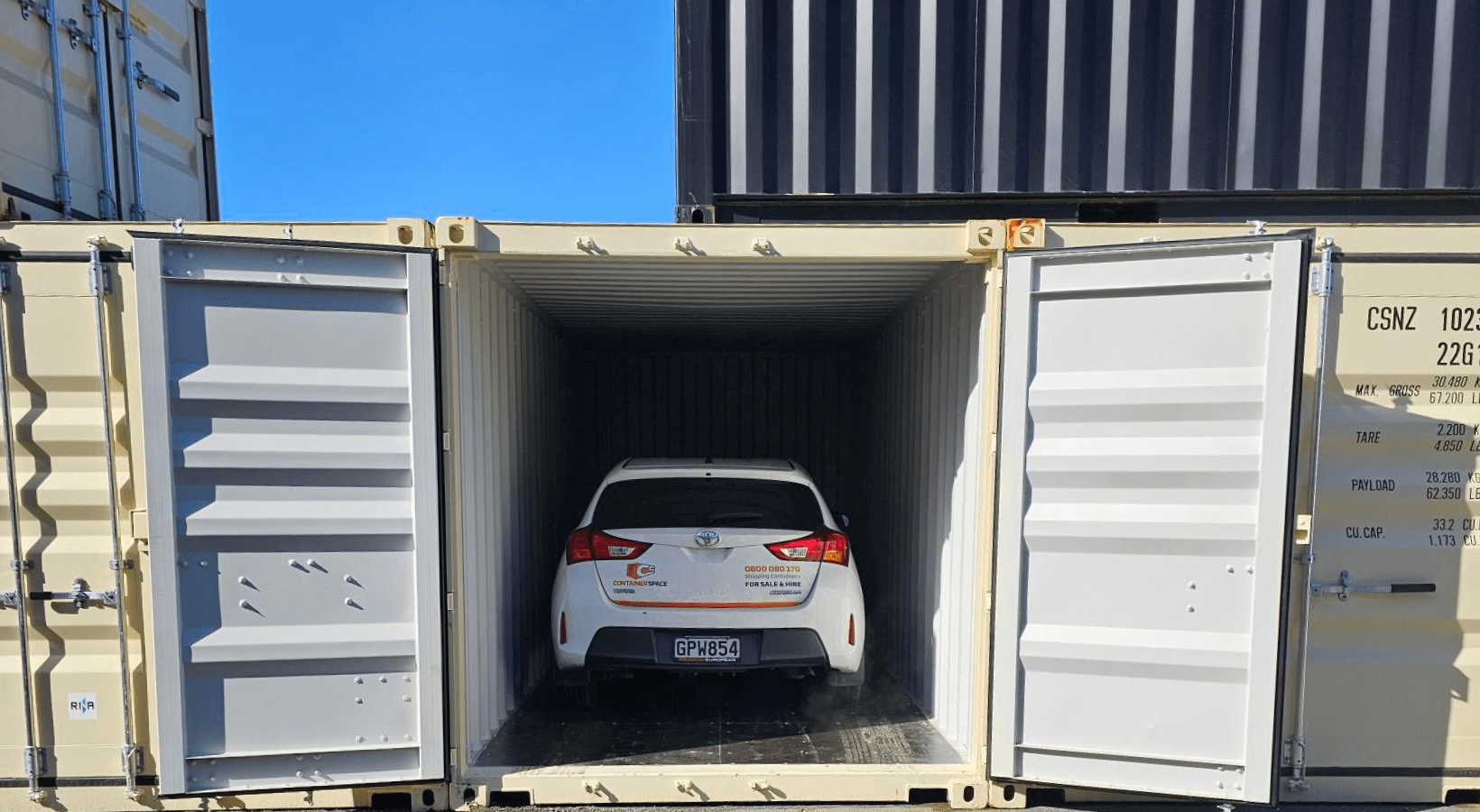 A white car is parked inside an open shipping container. The container is beige and located in an industrial area with other containers stacked nearby. The cars rear is visible with minor branding on it.