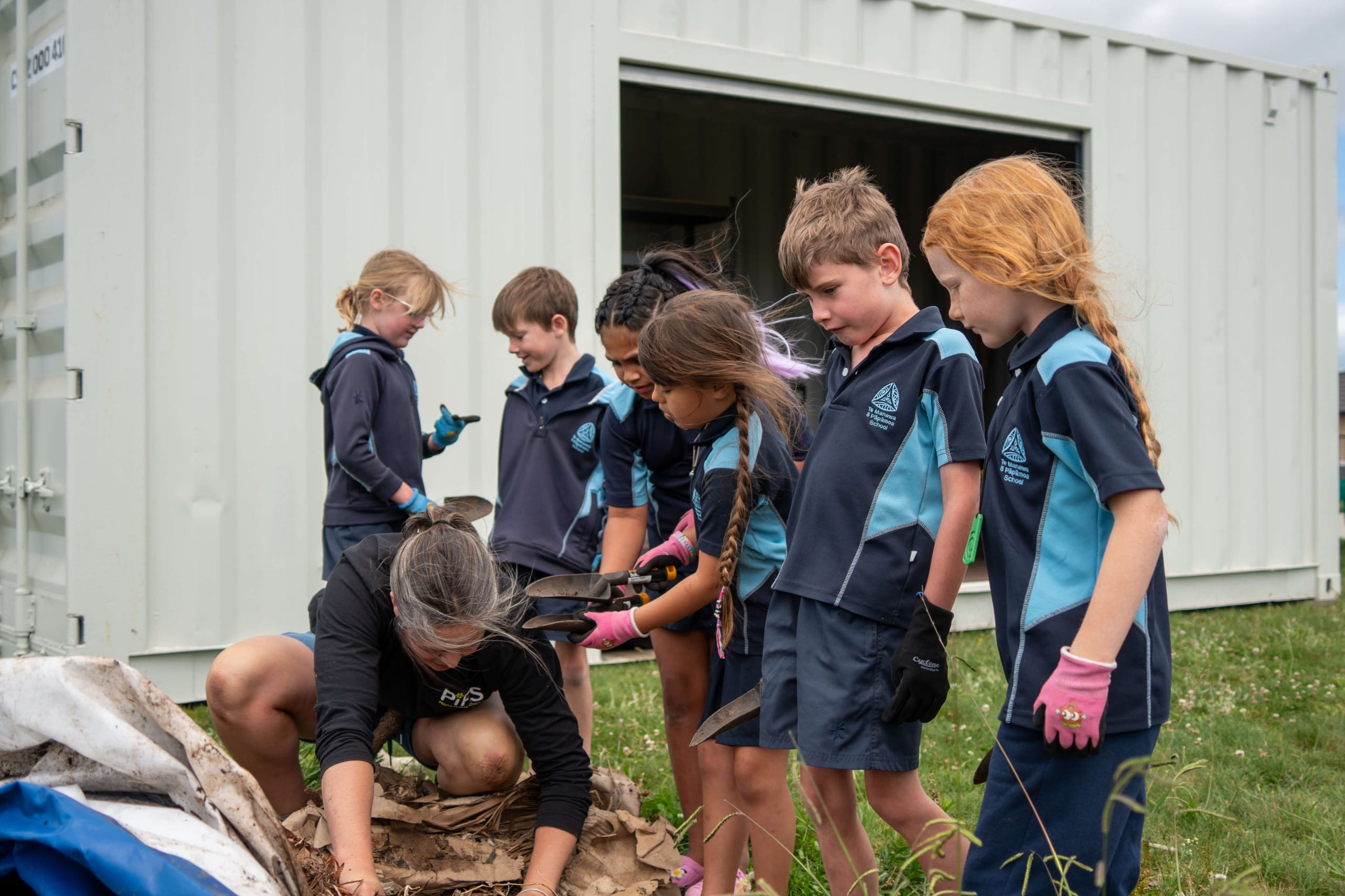 A group of children in school uniforms gather around a woman showing them something by a white shipping container, repurposed as a gardening shed. Some kids wear gloves, hinting at their outdoor gardening activity.