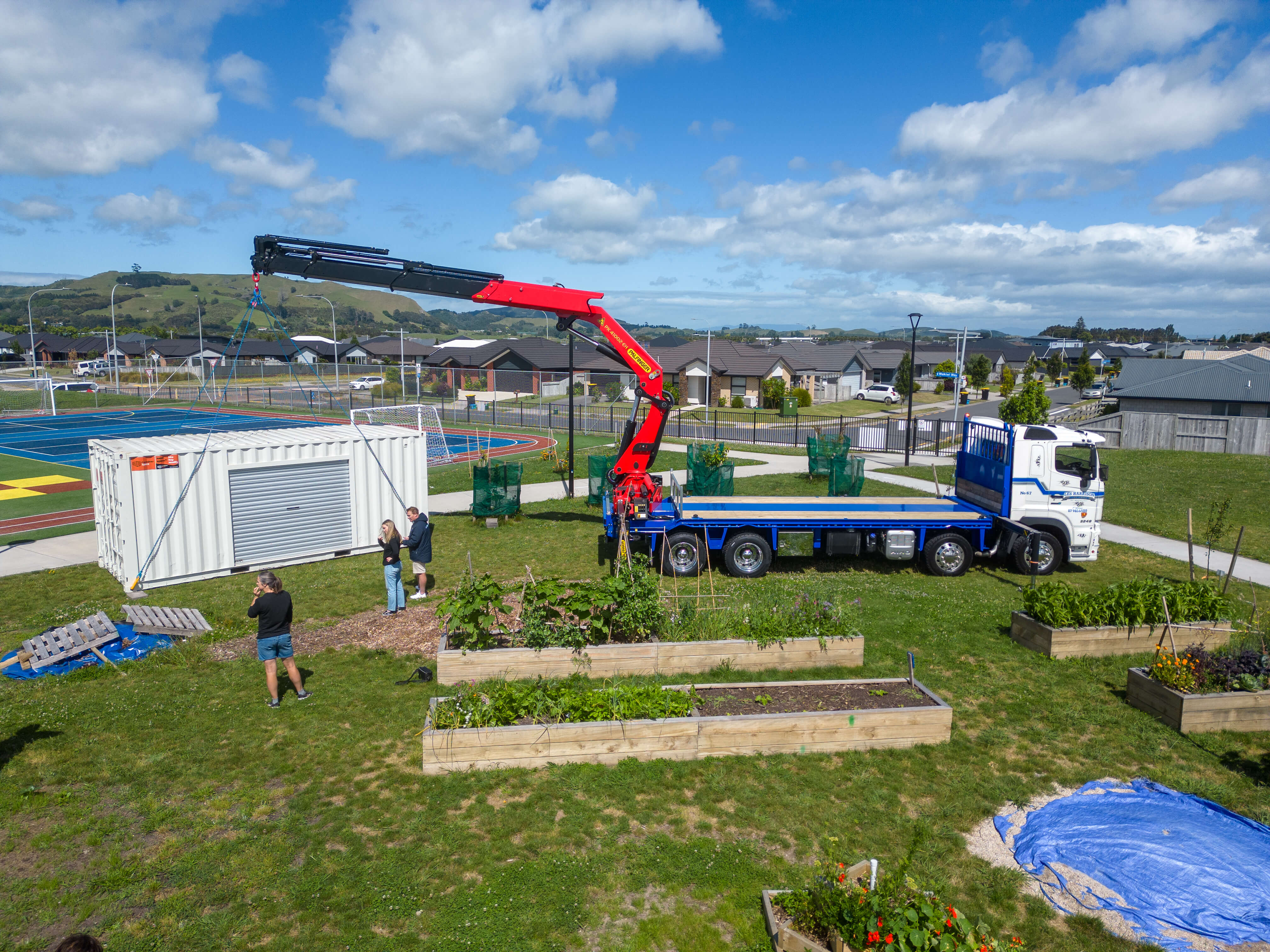 A crane truck carefully unloads a large white shipping container onto the grassy expanse of a community garden, destined to become a gardening shed. Several people guide the process, surrounded by raised garden beds with a colorful sports court and residential area in the backdrop.