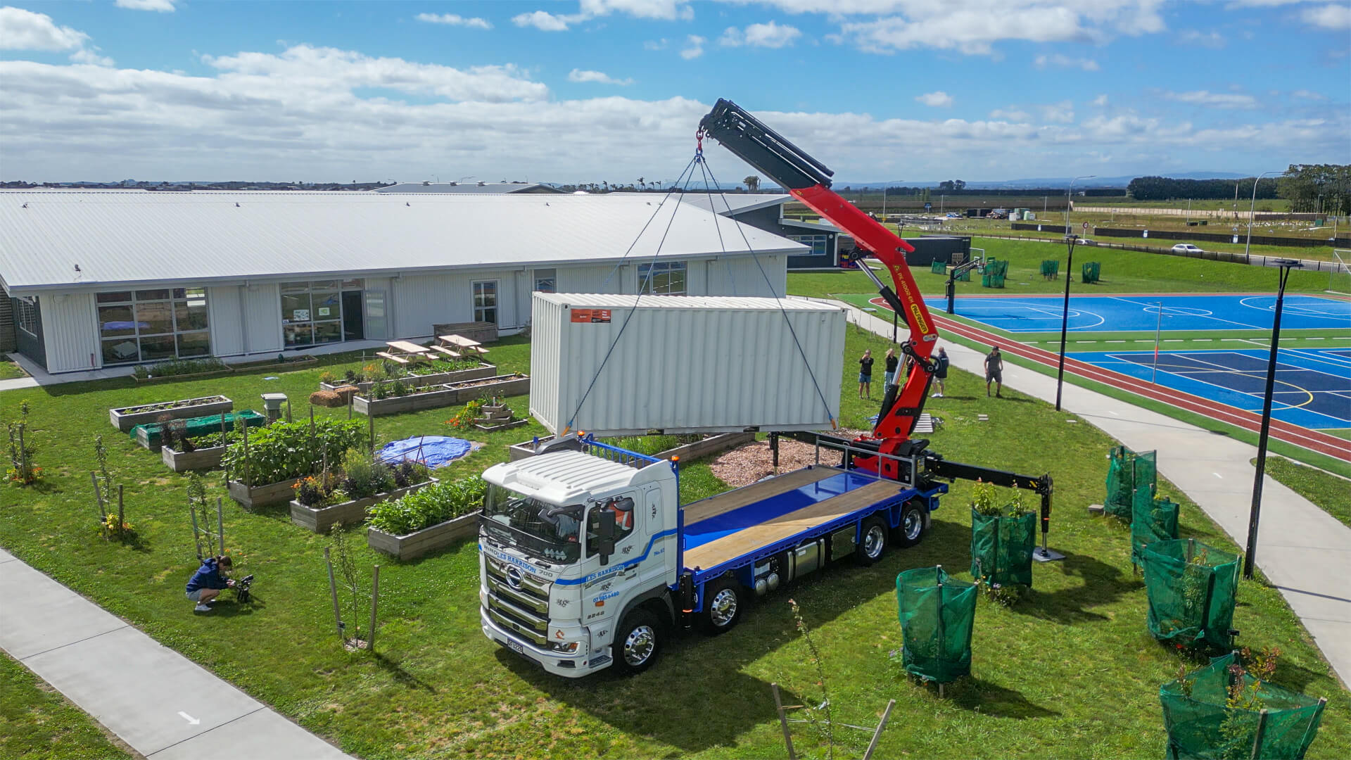 A truck with a mounted crane lifts a large white shipping container onto a grassy area beside the building, transforming it into an innovative gardening shed. Nearby, garden plots flourish, and young trees sway gently near the blue sports court under the partly cloudy sky.