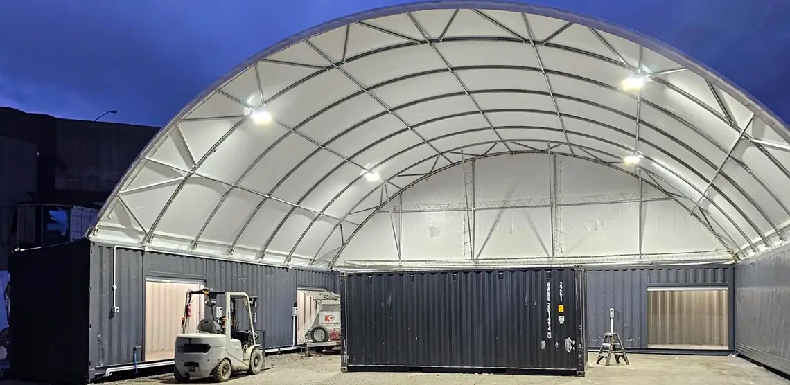 A large, white, tent-like industrial structure with tall, curved ceilings covers several shipping containers available for sale and hire on a gravel surface. A forklift is parked nearby, and bright lights illuminate the setup. The sky is dark, suggesting early evening.