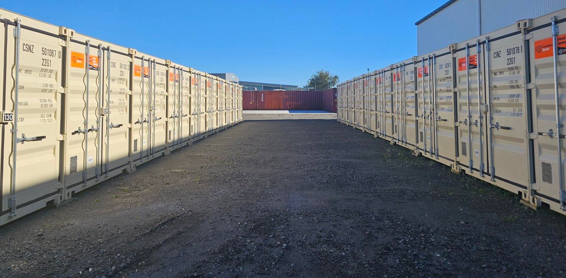 A row of white shipping containers for sale and hire lines each side of a gravel path under a clear blue sky. A red container is visible at the end of the path, with part of a building and some trees in the background.