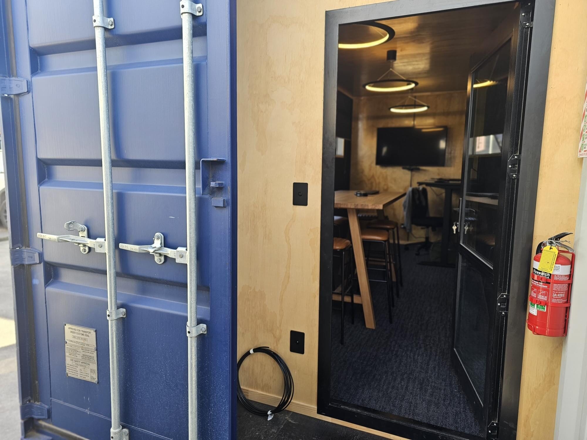 A blue shipping container, available for sale and hire, is transformed into an office featuring wooden interiors, a high table with stools, hanging lights, and a wall-mounted TV. The container door is open, revealing a fire extinguisher on the right.